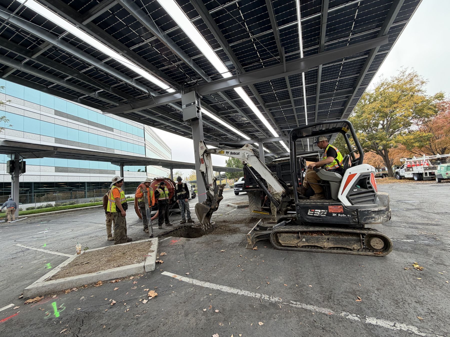 Apex crew excavating at a solar carport column with a mini excavator under the canopy