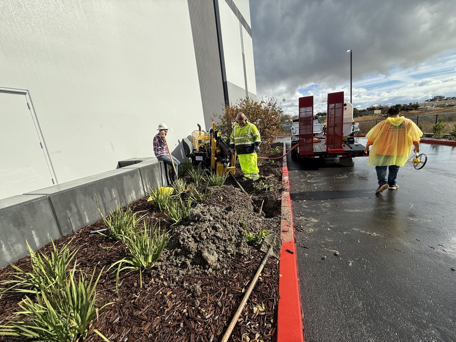 Crew running a compact drill in a planter strip between a building wall and a parking lot curb